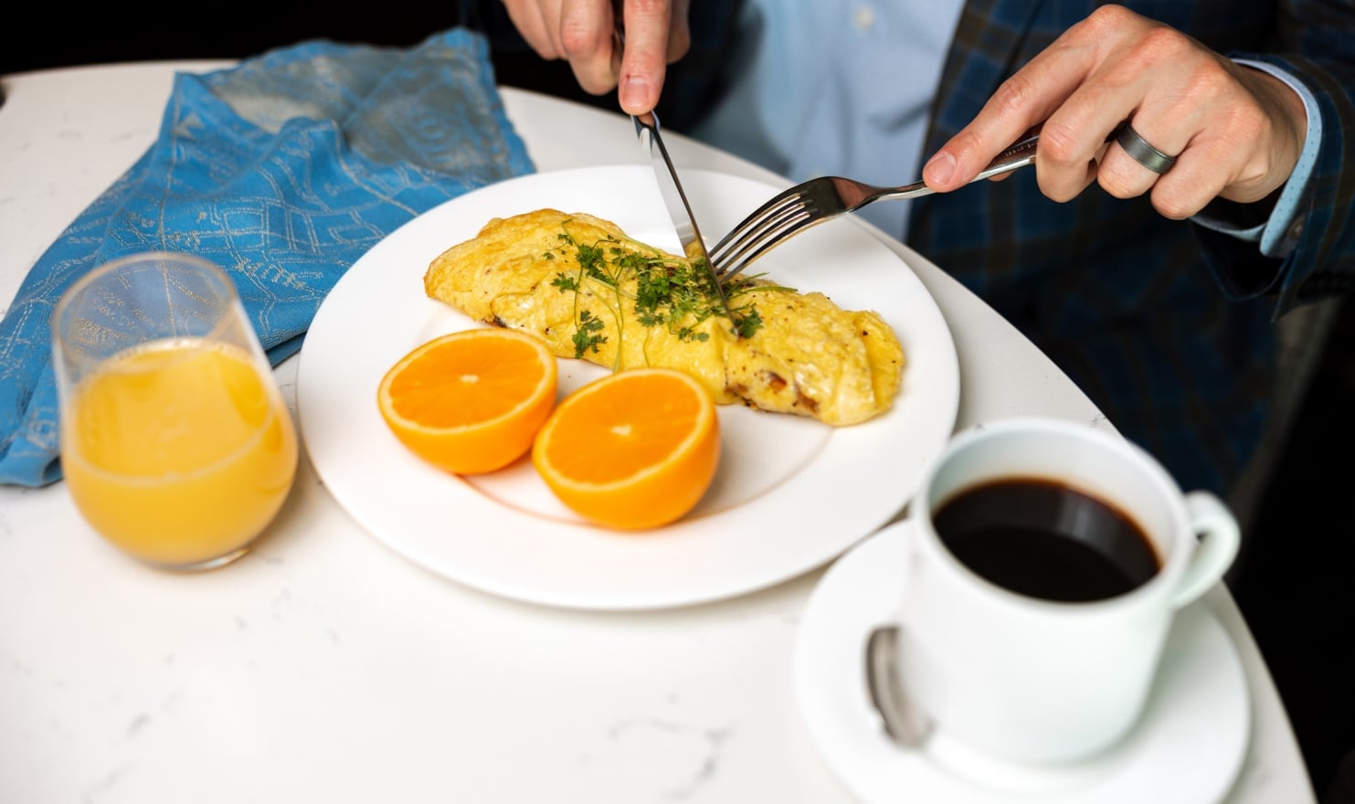 A person's hands, wearing a ring, cut into a herb-garnished omelet alongside orange halves, orange juice, and black coffee on a white table.