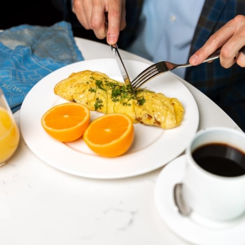 A person's hands, wearing a ring, cut into a herb-garnished omelet alongside orange halves, orange juice, and black coffee on a white table.