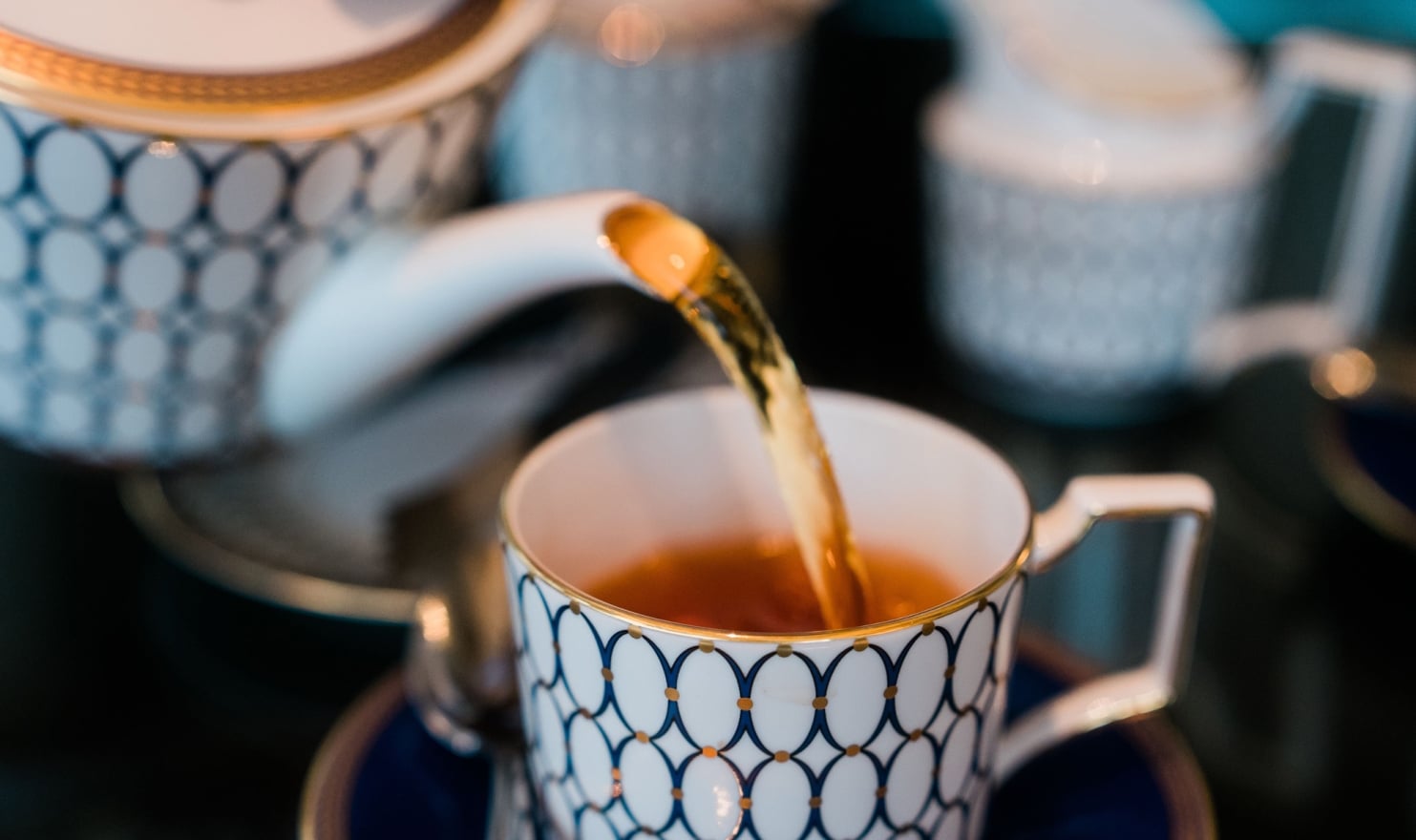 Tea is poured from a white and blue patterned teapot into a matching teacup. The elegant tea set sits on a dark surface, with other blurred components in the background.