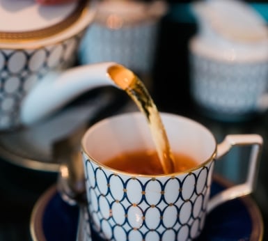 Tea is poured from a white and blue patterned teapot into a matching teacup. The elegant tea set sits on a dark surface, with other blurred components in the background.