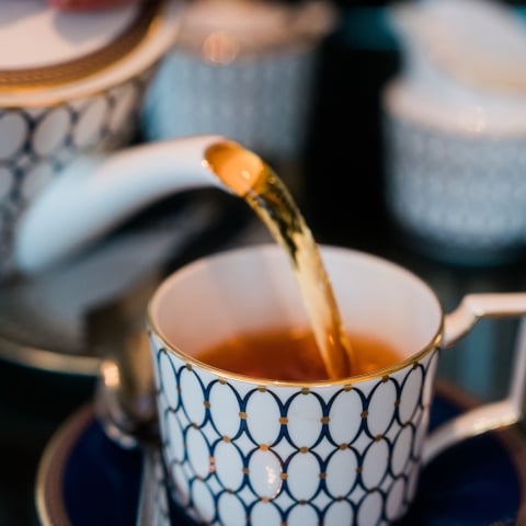 Tea is poured from a white and blue patterned teapot into a matching teacup. The elegant tea set sits on a dark surface, with other blurred components in the background.