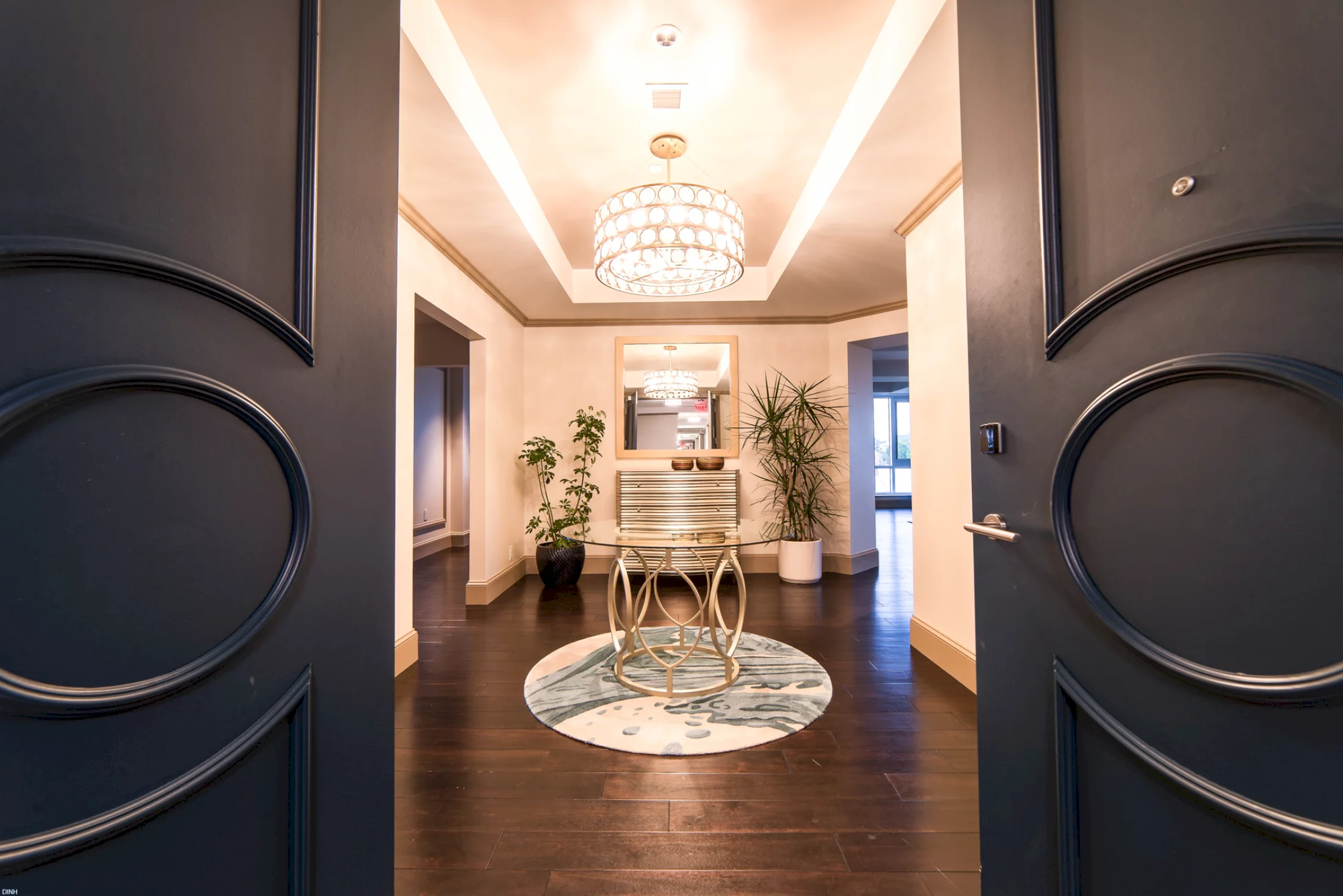 Open dark gray doors reveal a luxurious entryway with dark wood floors. A glass-top table on a round rug, flanked by plants, sits under a decorative chandelier.