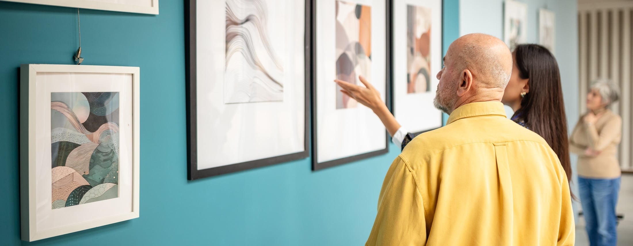 A bald man in a yellow shirt and a woman view abstract art on a teal wall in a gallery, with the woman gesturing towards a painting.