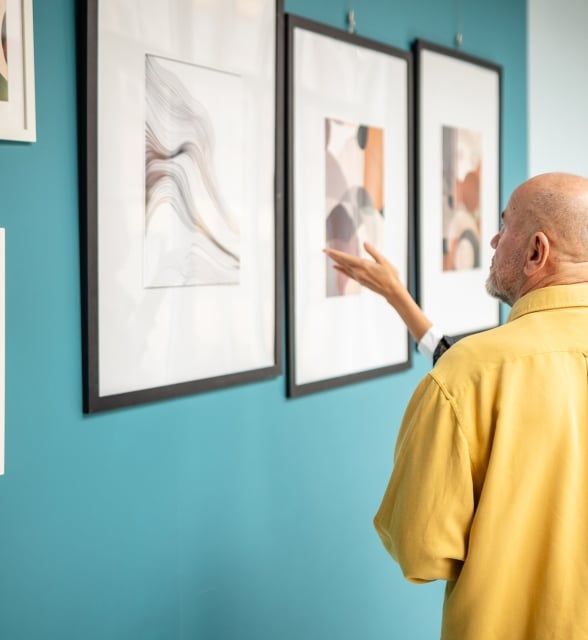 A bald man in a yellow shirt and a woman view abstract art on a teal wall in a gallery, with the woman gesturing towards a painting.