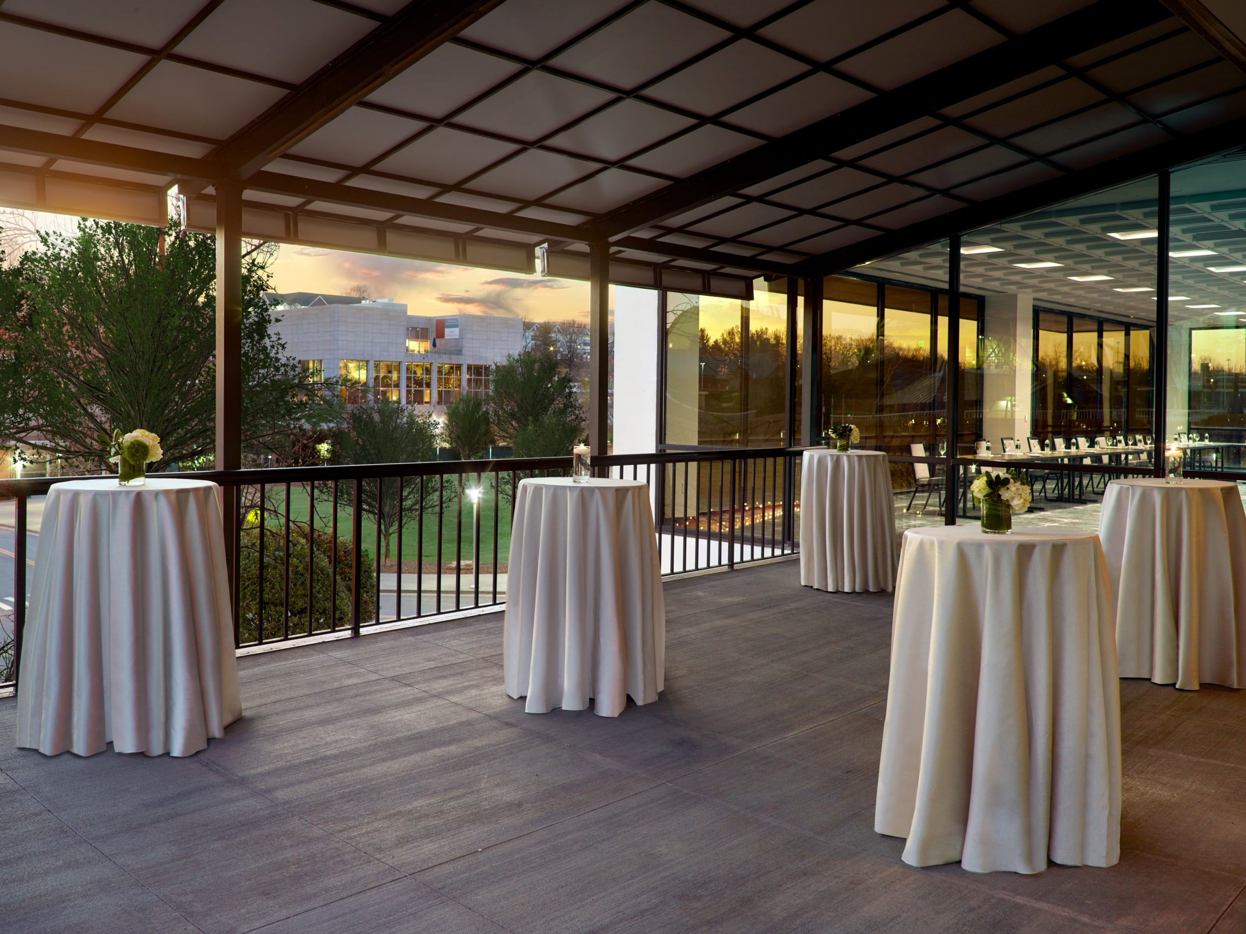 White-draped cocktail tables are arranged on a covered outdoor patio. It overlooks a park and modern buildings at sunset, next to an indoor event space through glass walls.