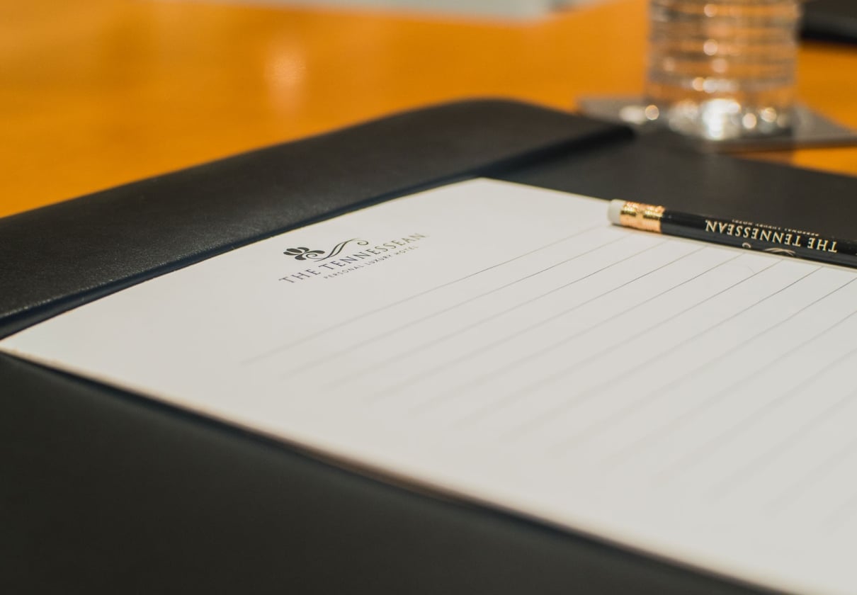 Lined white notepad and black pencil inscribed with "THE TENNESSEAN PERSONAL LUXURY HOTEL" rest on a black folio, ready for use on a warm wooden table, with a water glass in the background.