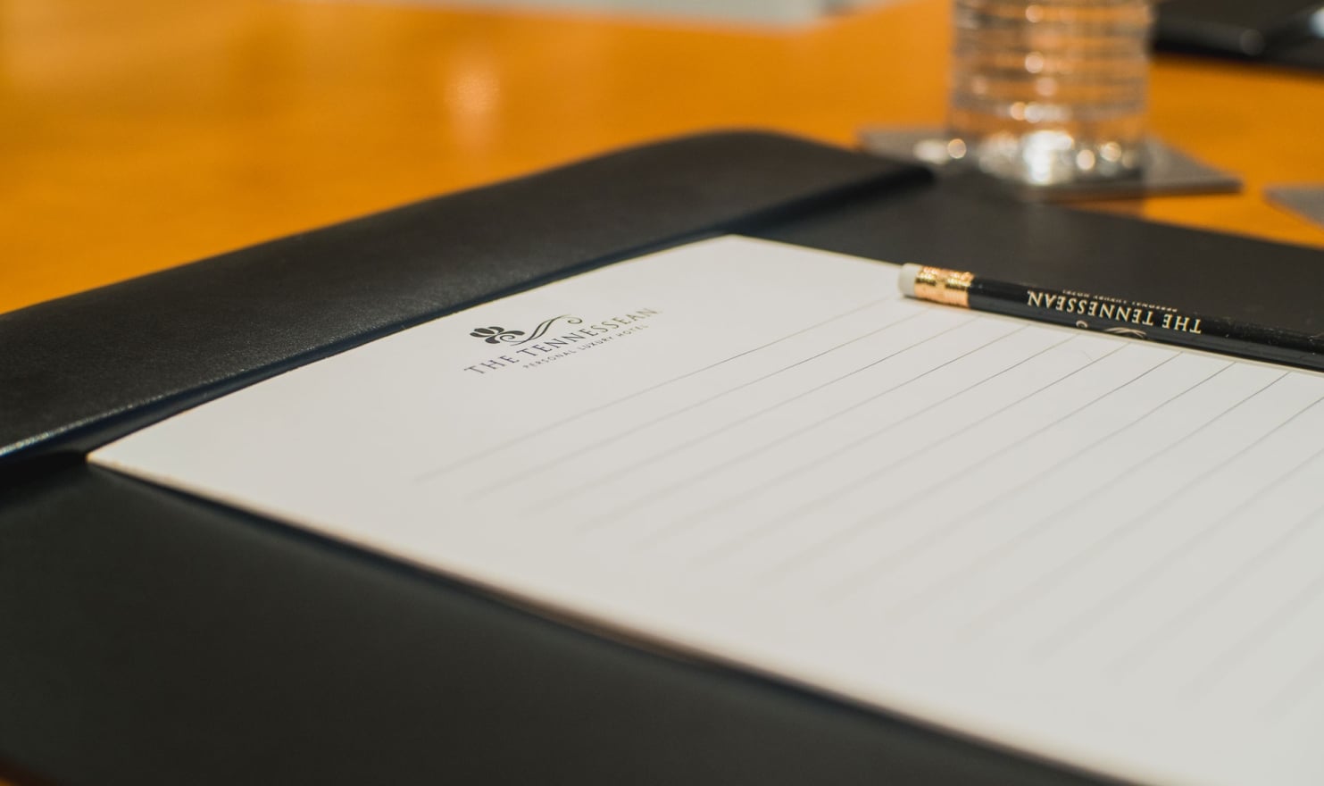 Lined white notepad and black pencil inscribed with "THE TENNESSEAN PERSONAL LUXURY HOTEL" rest on a black folio, ready for use on a warm wooden table, with a water glass in the background.