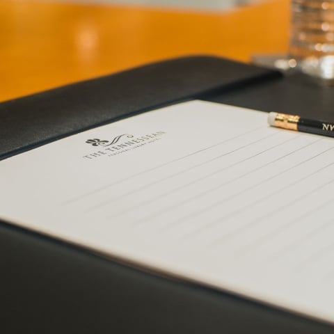Lined white notepad and black pencil inscribed with "THE TENNESSEAN PERSONAL LUXURY HOTEL" rest on a black folio, ready for use on a warm wooden table, with a water glass in the background.
