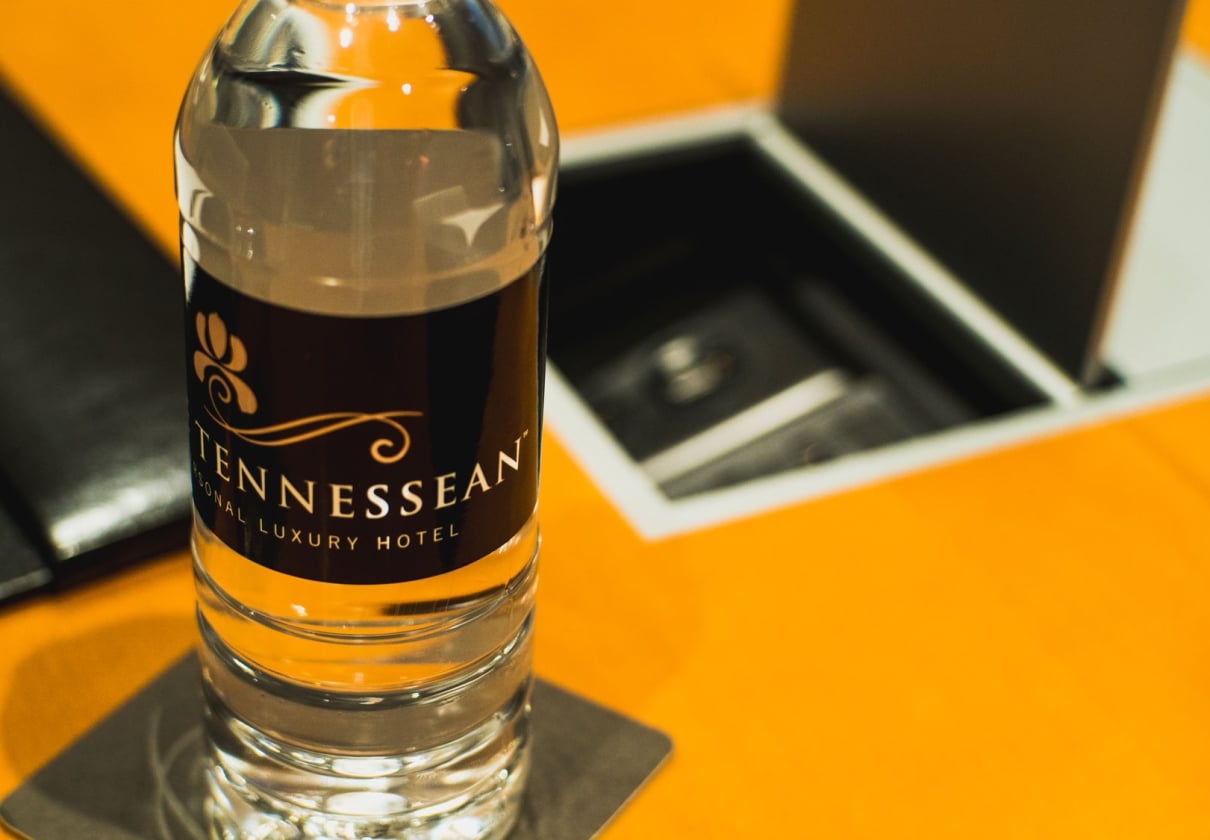 A clear water bottle with a "TENNESSEAN SONAL LUXURY HOTEL" label stands on a coaster, positioned on a bright orange surface next to an open, dark compartment.