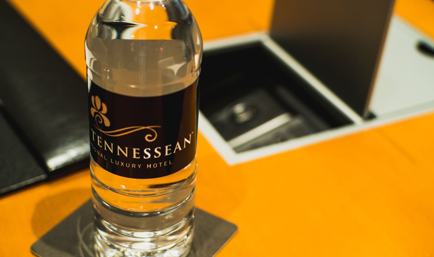 A clear water bottle with a "TENNESSEAN SONAL LUXURY HOTEL" label stands on a coaster, positioned on a bright orange surface next to an open, dark compartment.