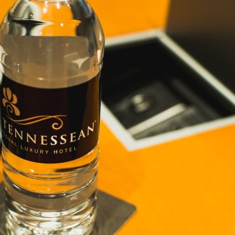 A clear water bottle with a "TENNESSEAN SONAL LUXURY HOTEL" label stands on a coaster, positioned on a bright orange surface next to an open, dark compartment.