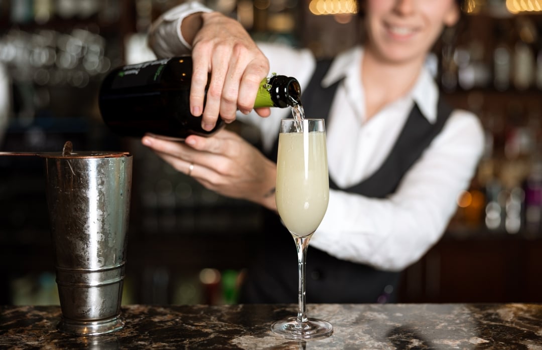 A bartender pours from a dark bottle into a champagne flute with a yellow drink, on a speckled bar counter beside a metal shaker, in a blurred bar setting.