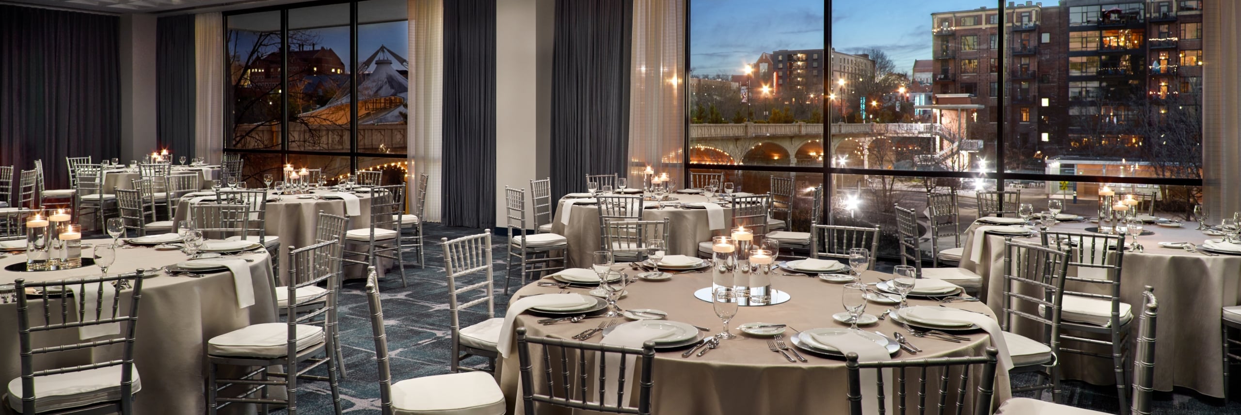 Round tables are set with place settings and candles in a ballroom overlooking a city street and illuminated bridge at dusk through large windows.
