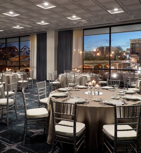 Round tables are set with place settings and candles in a ballroom overlooking a city street and illuminated bridge at dusk through large windows.