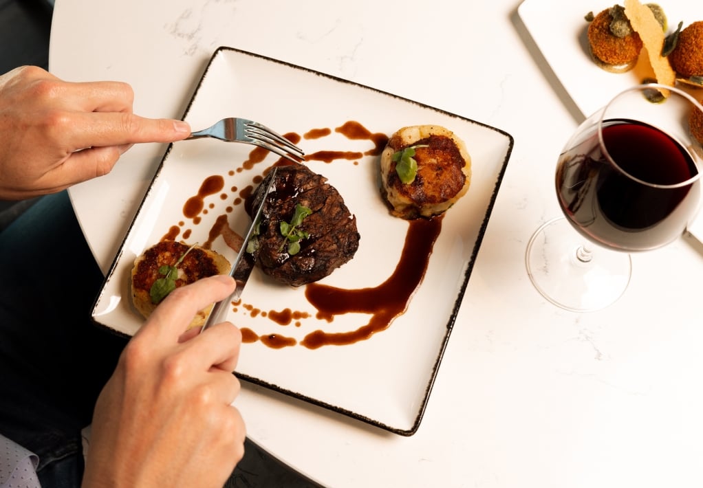 Hands are cutting a steak and potato cakes on a white plate with brown sauce, alongside a glass of red wine on a marble table.