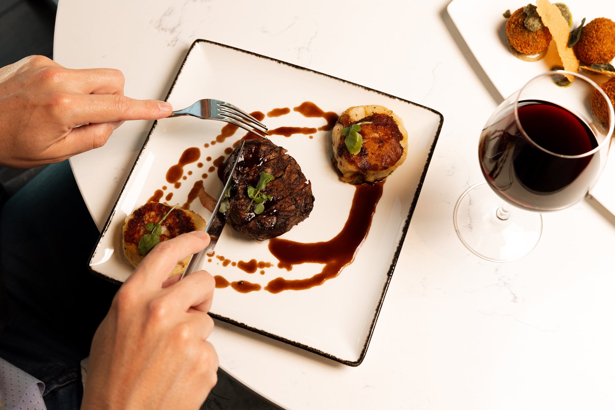 Hands are cutting a steak and potato cakes on a white plate with brown sauce, alongside a glass of red wine on a marble table.
