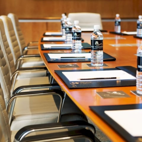 A polished conference table is prepared with "THE TENNESSEE PERSONAL LUXURY" bottled water, notepads, and pencils. Empty cream-colored chairs surround the table in a meeting room.