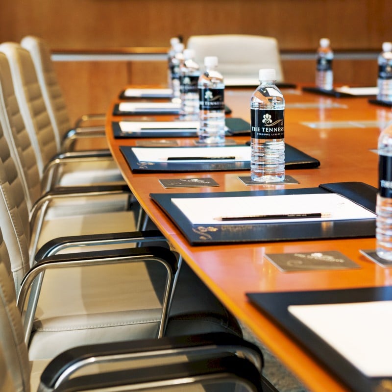 A polished conference table is prepared with "THE TENNESSEE PERSONAL LUXURY" bottled water, notepads, and pencils. Empty cream-colored chairs surround the table in a meeting room.