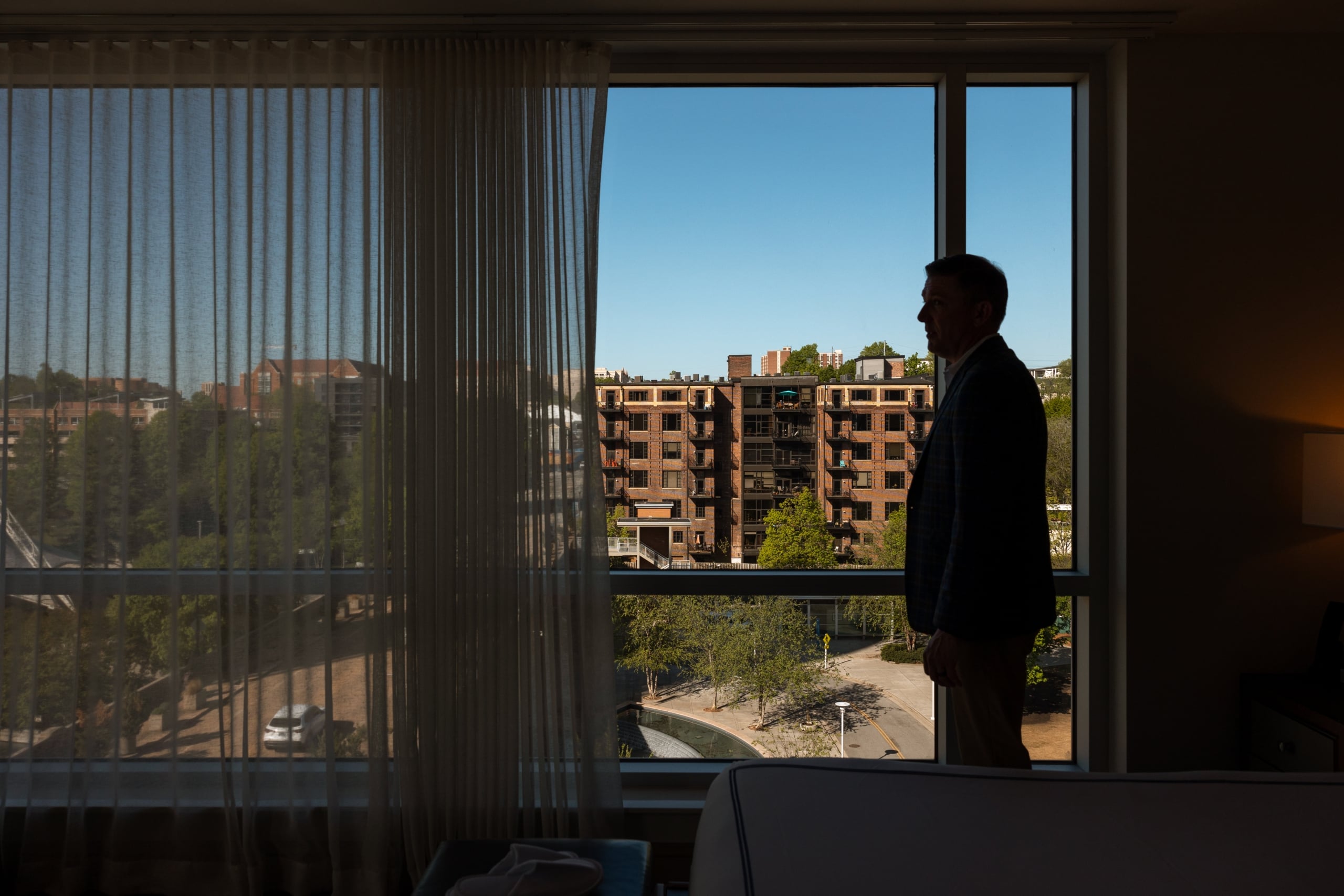 A silhouetted man gazes out a large window at a sunny urban landscape featuring multi-story brick buildings, green trees, and roads under a clear blue sky.