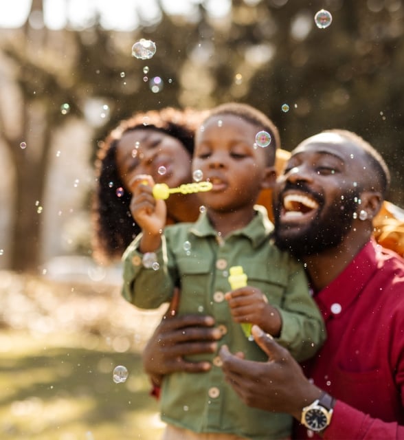 A young Black boy blows bubbles from a yellow wand, held by his smiling parents who are laughing, surrounded by floating bubbles outdoors on a sunny day.