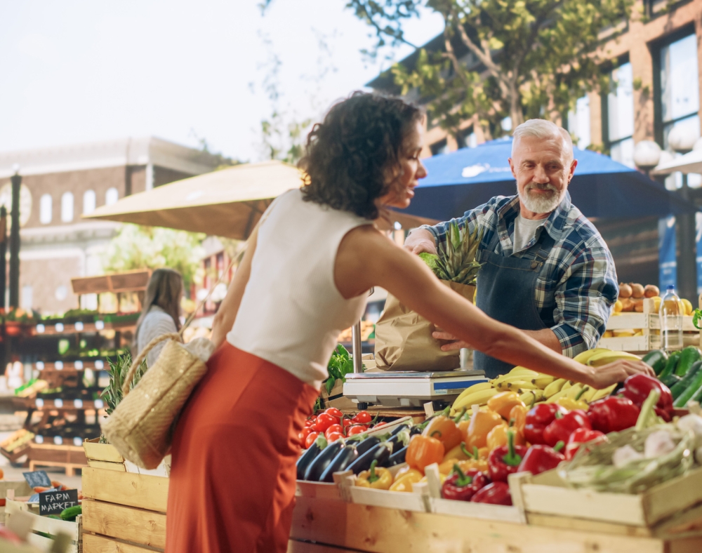 A woman chooses produce while a vendor bags items from a stall laden with fruits and vegetables at a sunny outdoor "FARM MARKET".