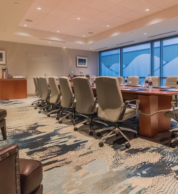 A modern conference room features a long wooden table surrounded by numerous beige office chairs. It has a blue and tan patterned carpet, large windows, and a refreshment counter. Text: Acqua Panna
