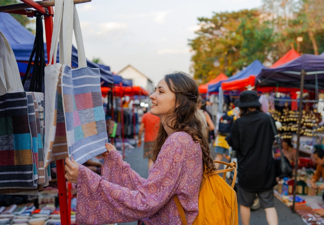 A young woman inspects patterned tote bags hanging from a stall at a lively outdoor market with numerous stalls under red and blue tarps.