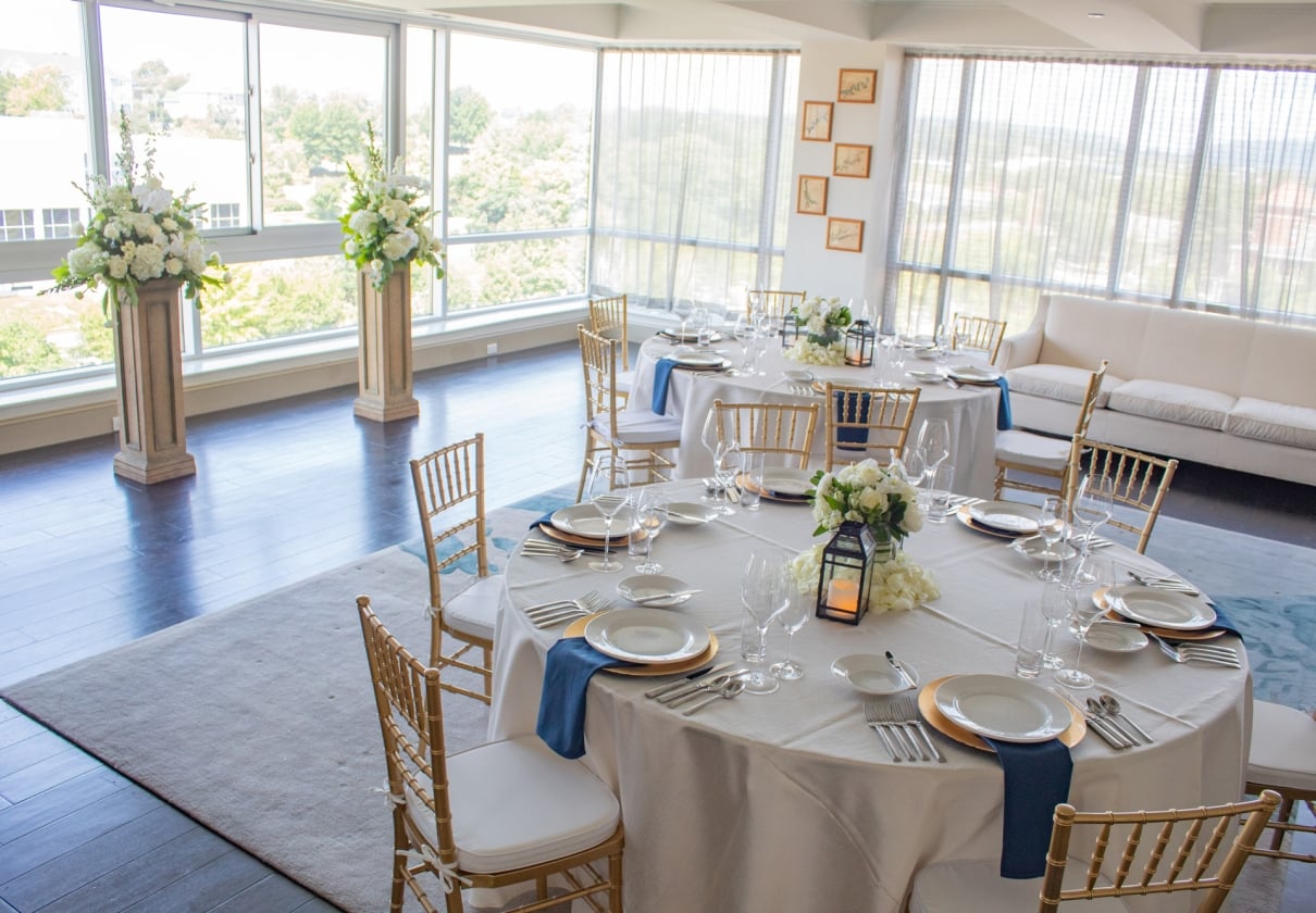 Elegant round tables are set with white linens, gold chargers, and blue napkins, surrounded by gold chairs, in a bright, modern room with large windows and floral pedestals.
