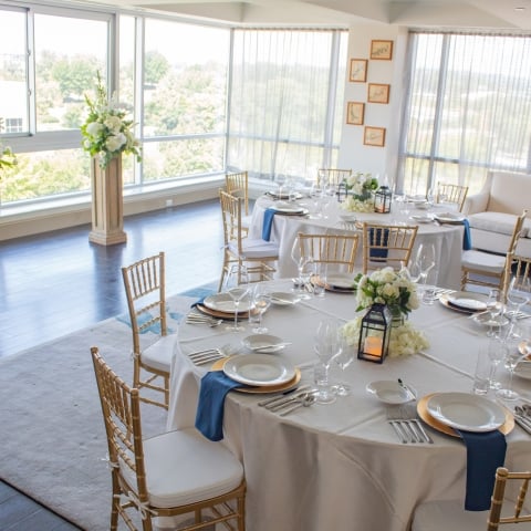 Elegant round tables are set with white linens, gold chargers, and blue napkins, surrounded by gold chairs, in a bright, modern room with large windows and floral pedestals.