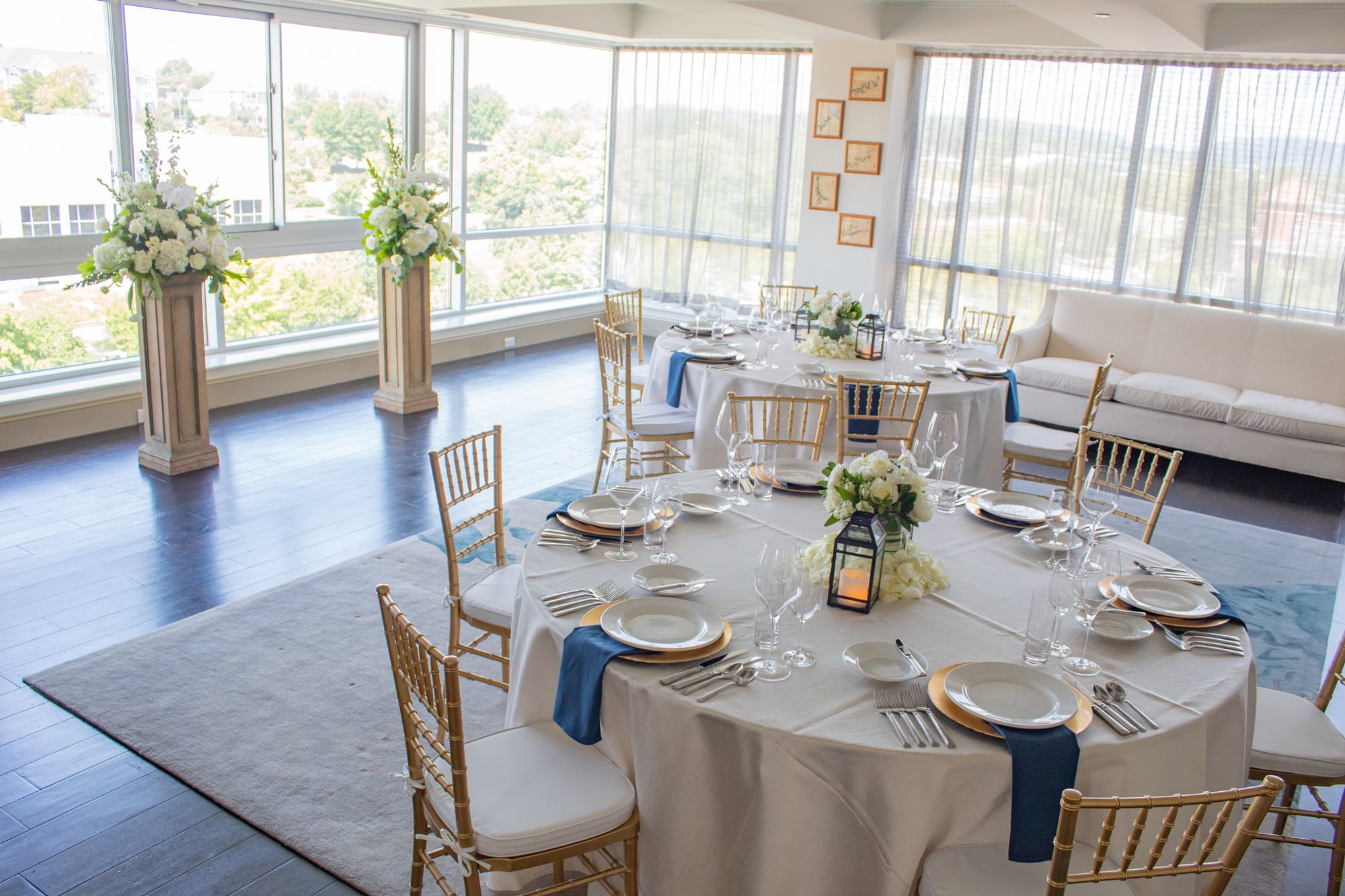 Elegant round tables are set with white linens, gold chargers, and blue napkins, surrounded by gold chairs, in a bright, modern room with large windows and floral pedestals.