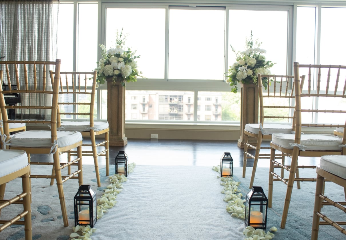 A wedding aisle, defined by a white runner, is flanked by gold chairs, black lanterns, and floral pedestals, leading to bright windows overlooking a city.