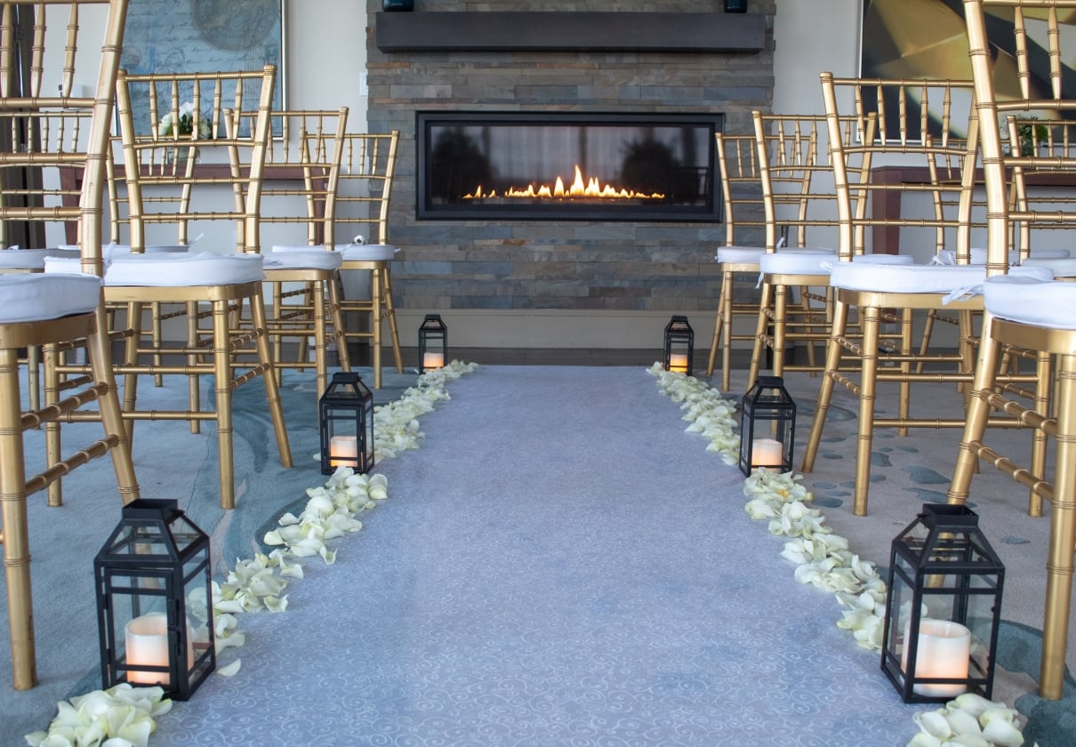 A wedding aisle leads towards a lit modern fireplace, flanked by gold bamboo-style chairs with white cushions, lanterns, and white rose petals in an indoor event space.
