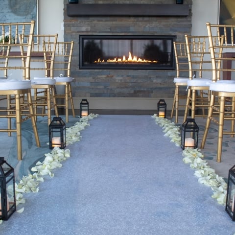 A wedding aisle leads towards a lit modern fireplace, flanked by gold bamboo-style chairs with white cushions, lanterns, and white rose petals in an indoor event space.