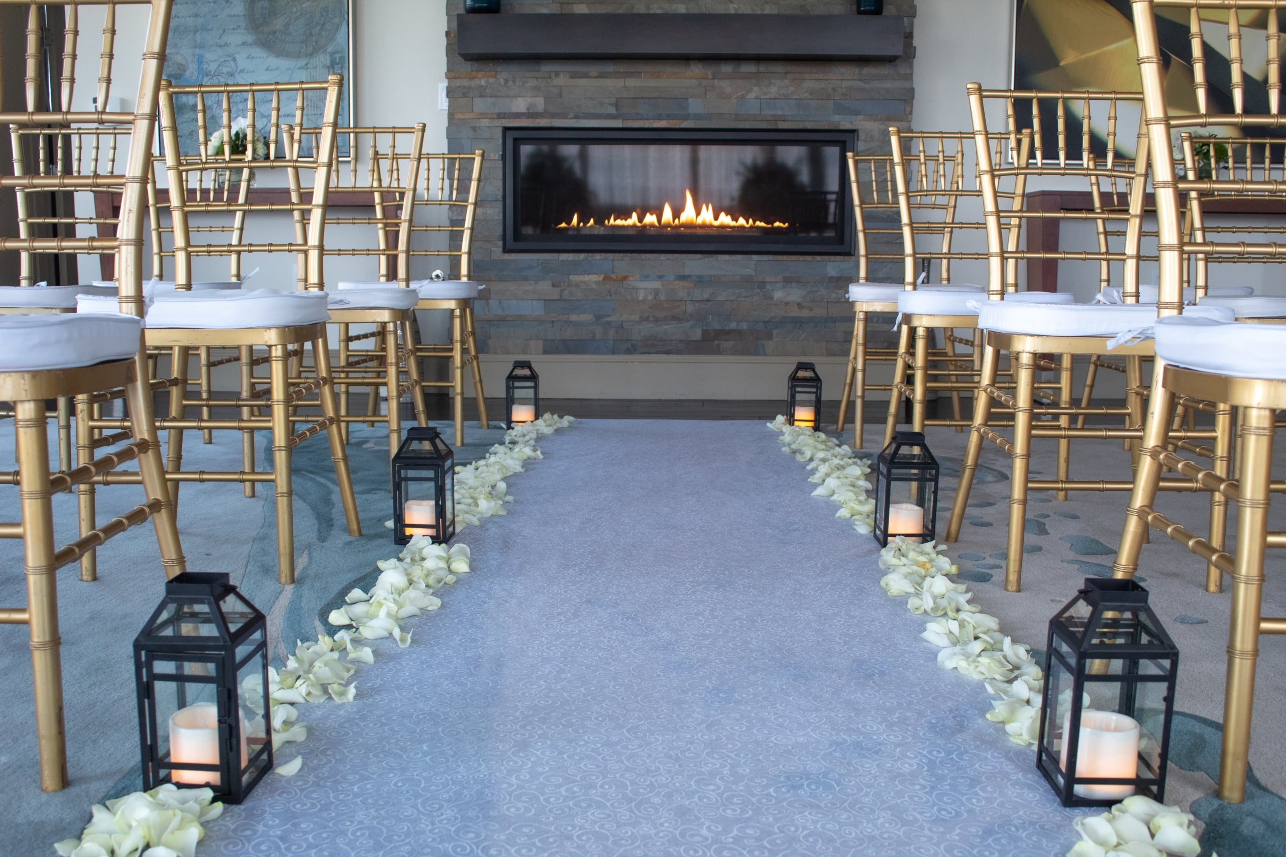 A wedding aisle leads towards a lit modern fireplace, flanked by gold bamboo-style chairs with white cushions, lanterns, and white rose petals in an indoor event space.