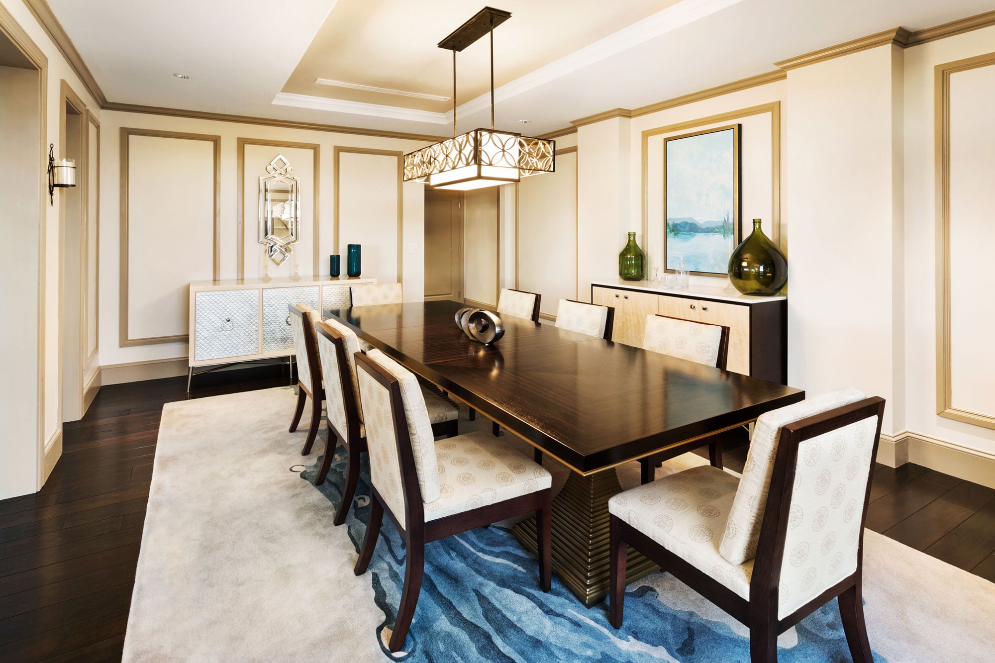 A long, dark wooden dining table, surrounded by eight cream chairs, anchors a luxurious room. A blue wavy rug, ornate sideboards, and a rectangular chandelier complete the opulent context.