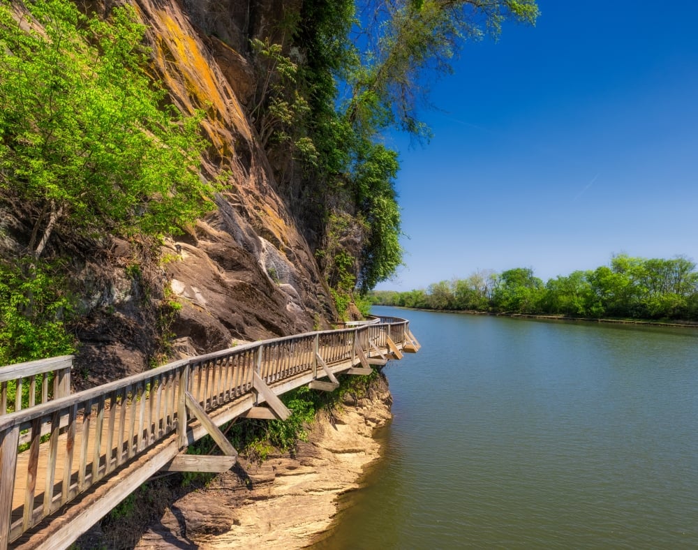 A wooden boardwalk curves along a green-vegetated rocky cliff, overlooking a wide river with trees on the opposite bank under a clear blue sky.