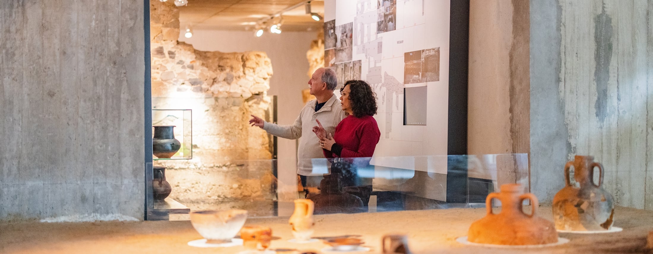 A couple discusses ancient pottery exhibits in a modern museum with exposed stone walls. The man gestures towards a display, surrounded by artifacts and informational panels.