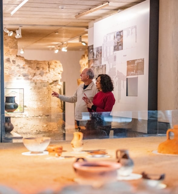A couple discusses ancient pottery exhibits in a modern museum with exposed stone walls. The man gestures towards a display, surrounded by artifacts and informational panels.