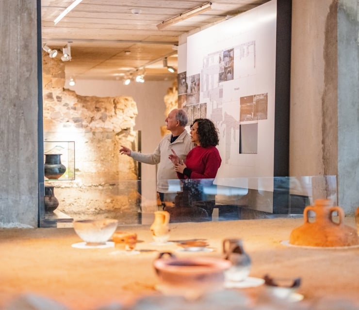 A couple discusses ancient pottery exhibits in a modern museum with exposed stone walls. The man gestures towards a display, surrounded by artifacts and informational panels.