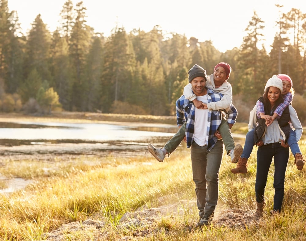 A smiling family walks through a sunny field beside a lake. The two adults give their children piggyback rides, with evergreen trees in the background.