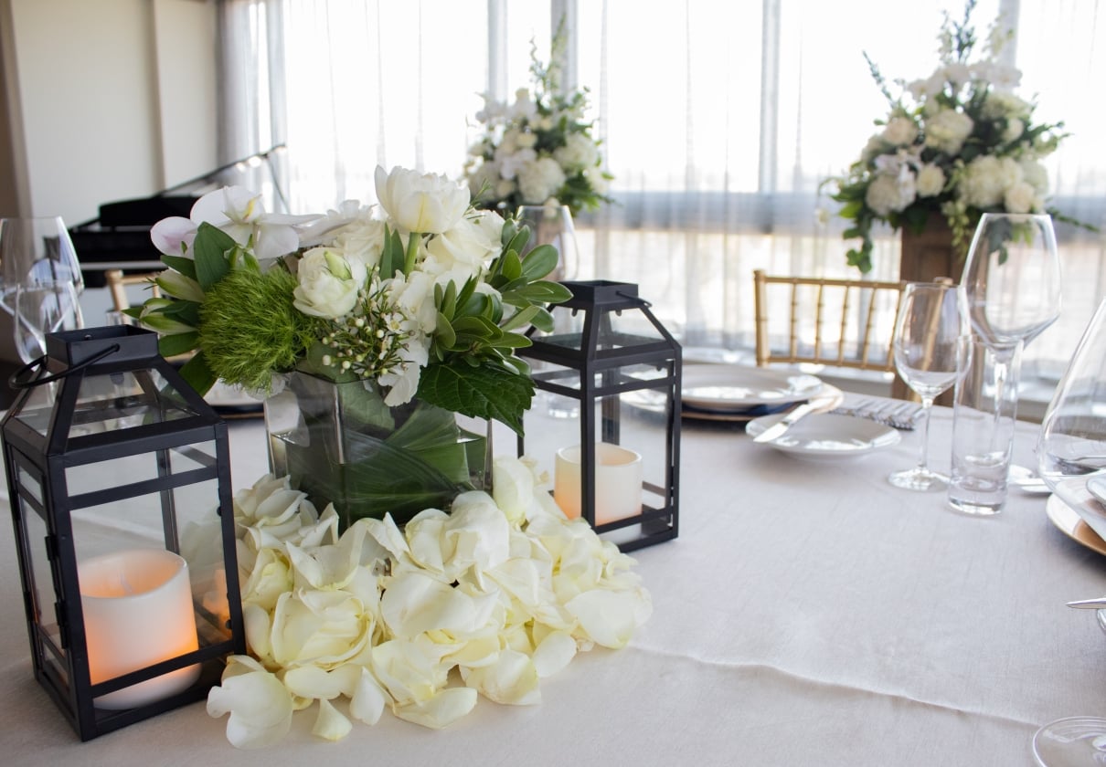 A formal dining table elegantly set with white rose floral centerpieces, lit lanterns, and gold chairs.