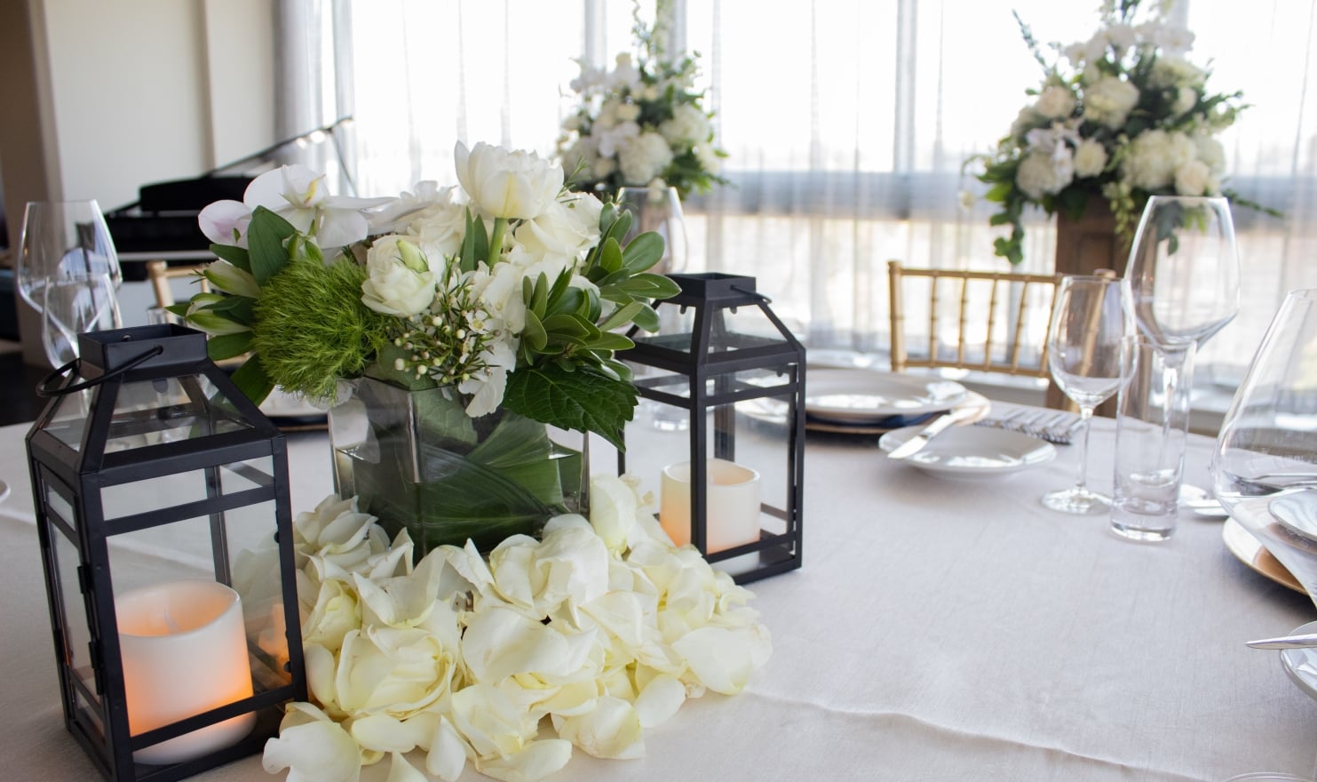 A formal dining table elegantly set with white rose floral centerpieces, lit lanterns, and gold chairs.