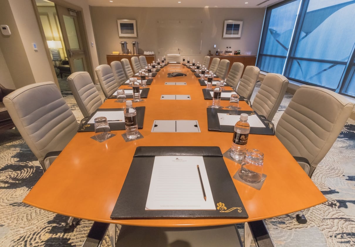 A long orange boardroom table is set with "THE TENNESSEAN HOTEL" water bottles, glasses, and notepads. Empty tan chairs, a patterned rug, and blue-tinted windows fill the room.