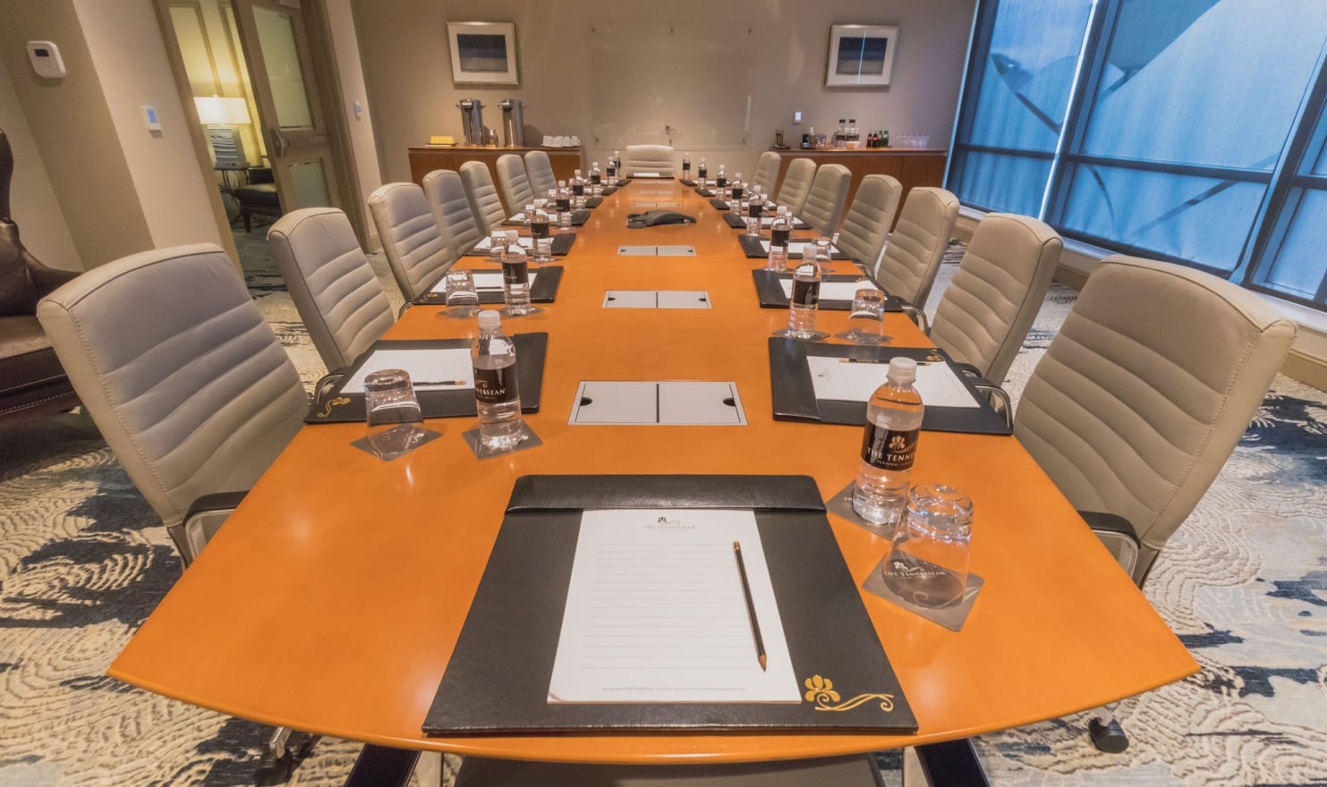 A long orange boardroom table is set with "THE TENNESSEAN HOTEL" water bottles, glasses, and notepads. Empty tan chairs, a patterned rug, and blue-tinted windows fill the room.