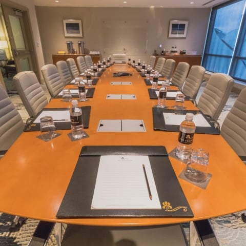 A long orange boardroom table is set with "THE TENNESSEAN HOTEL" water bottles, glasses, and notepads. Empty tan chairs, a patterned rug, and blue-tinted windows fill the room.