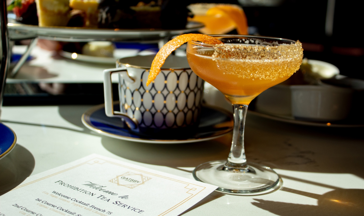An orange cocktail in a stemmed glass with a sugared rim and orange peel sits on a white table. A "PROHIBITION TEA SERVICE" menu, teacup, and tiered desserts are behind it.