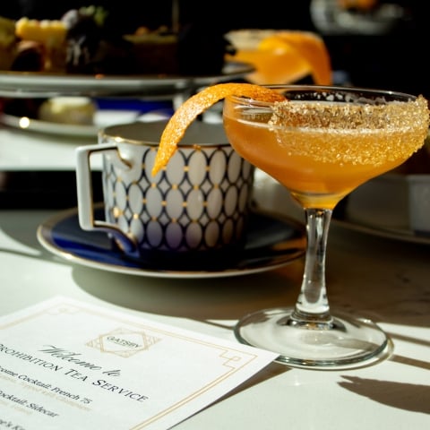 An orange cocktail in a stemmed glass with a sugared rim and orange peel sits on a white table. A "PROHIBITION TEA SERVICE" menu, teacup, and tiered desserts are behind it.