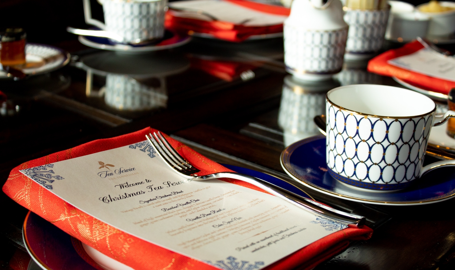 Multiple elegant tea settings, featuring white and blue patterned cups and saucers, are arranged on a dark, reflective table for a "Christmas Tea Service."