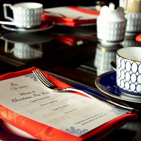 Multiple elegant tea settings, featuring white and blue patterned cups and saucers, are arranged on a dark, reflective table for a "Christmas Tea Service."