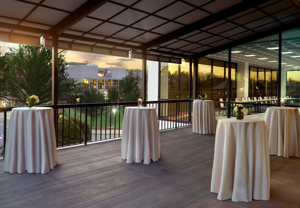 White draped high-top tables stand on a covered outdoor patio overlooking a building and trees at sunset, adjacent to an indoor event space with glass walls.